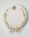 A set of gold celestial-themed hoop earrings with star, moon, and flower charms, displayed on a white circular stand against a light gray background.
