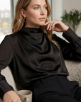 Woman wearing a black satin cowl neck blouse with long sleeves and elegant draped neckline.