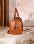 A brown leather handbag with a top handle and shoulder strap, styled on a textured hide rug next to an open magazine against a rustic wall and curtain backdrop.