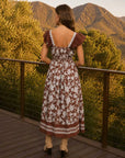 A woman stands on a wooden deck overlooking a mountain landscape at sunset, wearing a brown and white floral midi dress with ruffled sleeves and beige ankle boots.