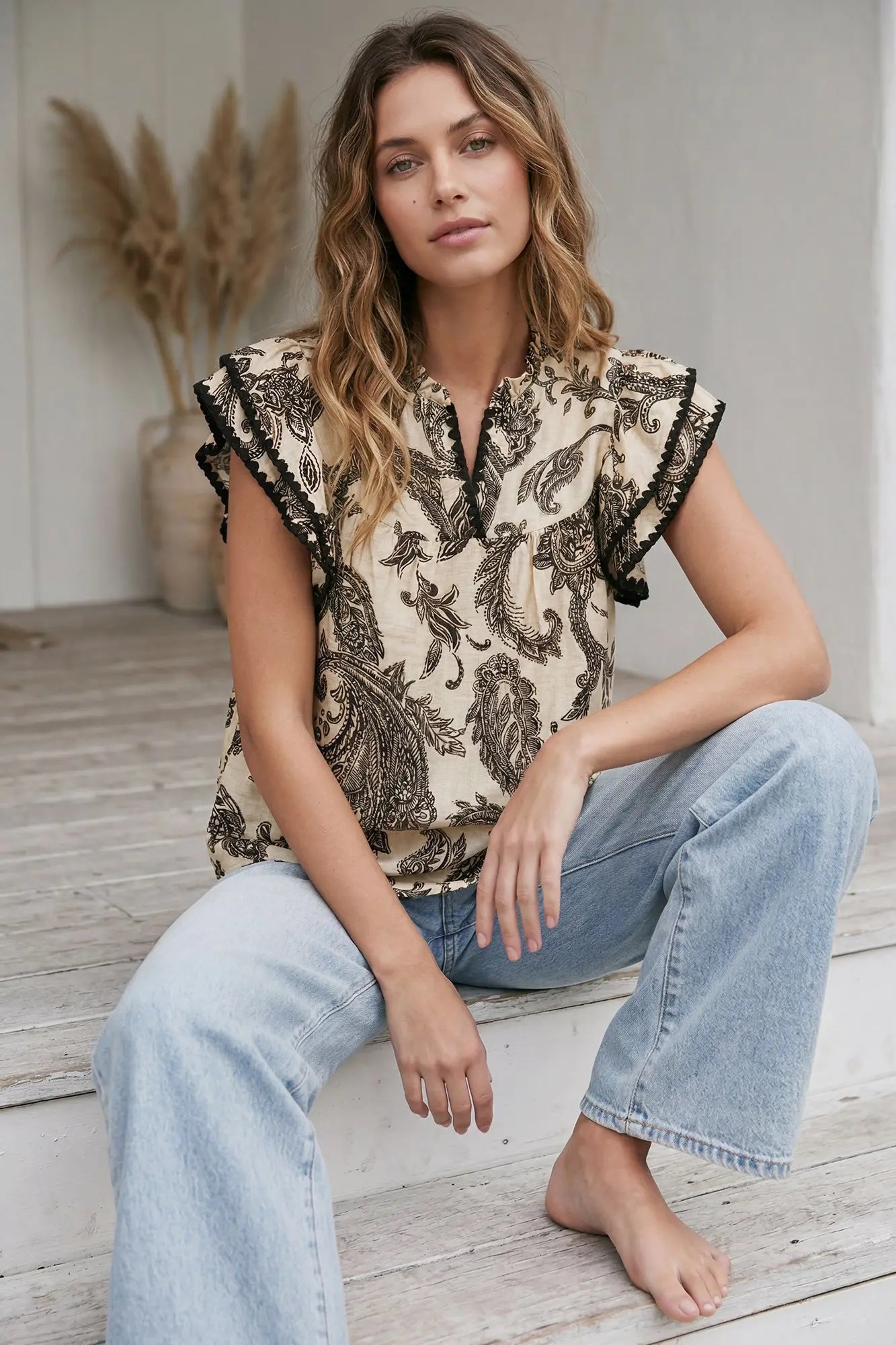 Woman wearing a cream and black paisley print woven top with ruffle sleeves, contrast trim, and a split neckline paired with light wash jeans.