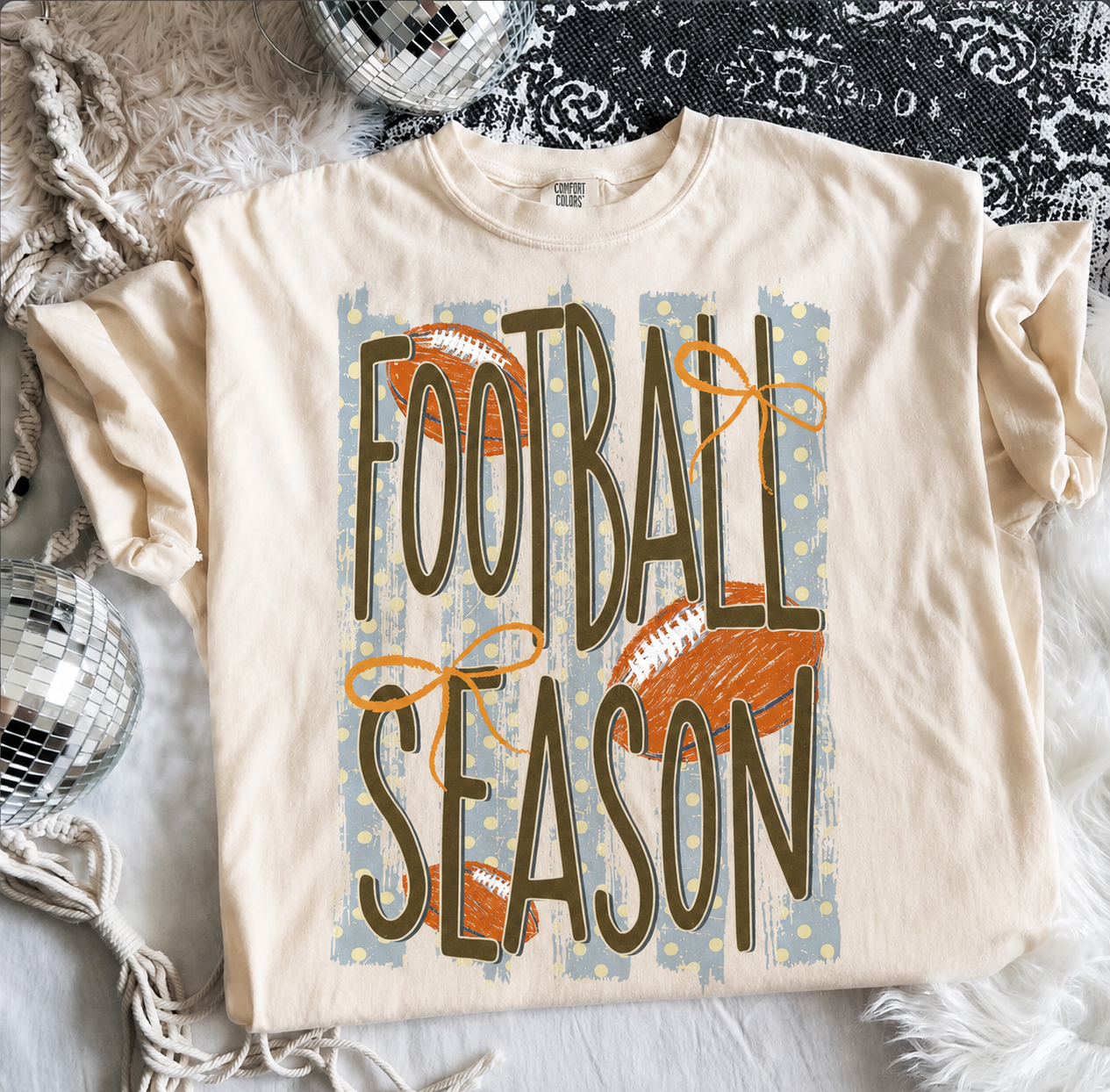 Cream Comfort Colors® Football Season graphic tee featuring vintage football artwork, distressed lettering, orange bows, and a relaxed unisex fit.