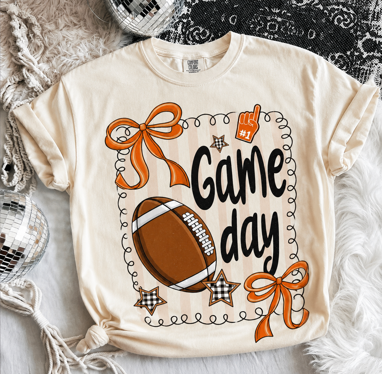 Cream Comfort Colors® Game Day graphic tee featuring a shimmering football design, orange bows, football artwork, and a relaxed unisex fit.