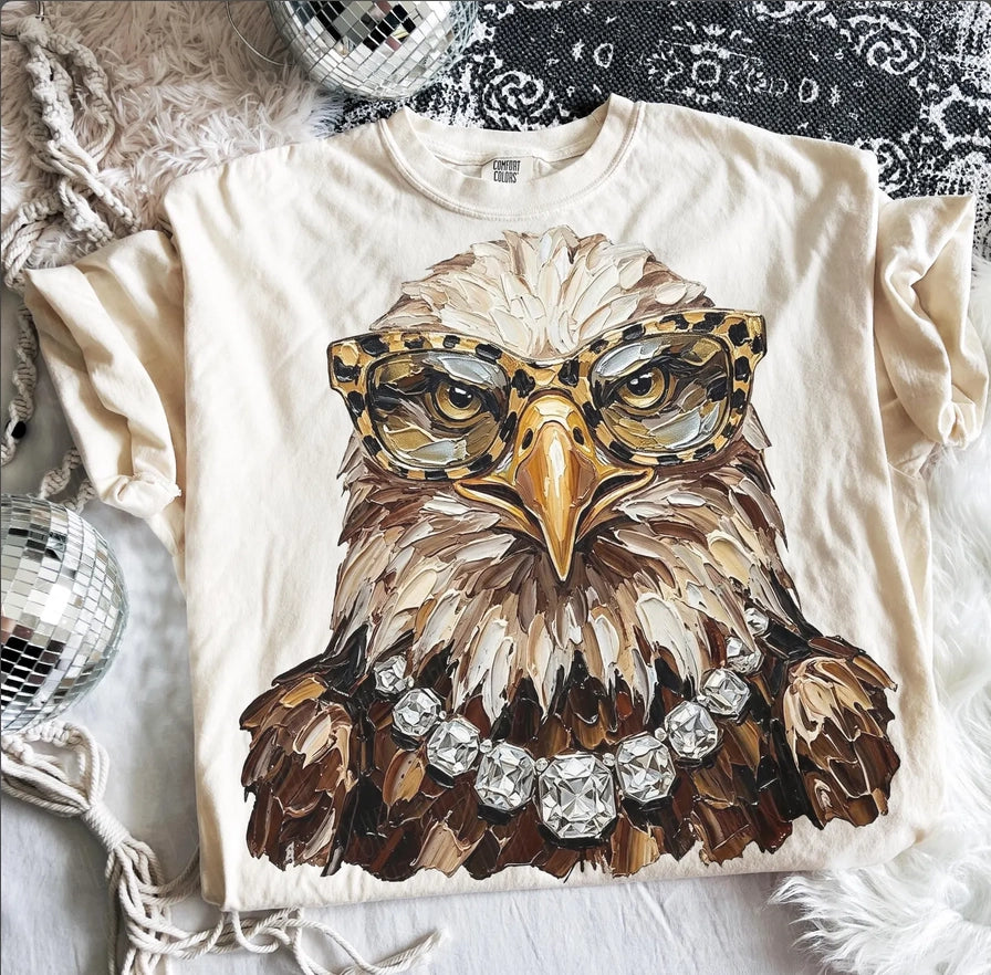 Woman’s ivory Comfort Colors graphic tee featuring a glam eagle wearing leopard print glasses and a rhinestone necklace.
