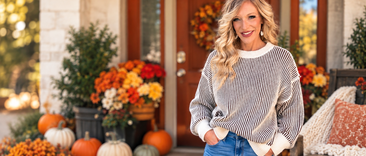 Halemi & Co. model wearing a cream and brown striped sweater with blue jeans on a cozy fall porch decorated with pumpkins and autumn flowers. Copy alt text