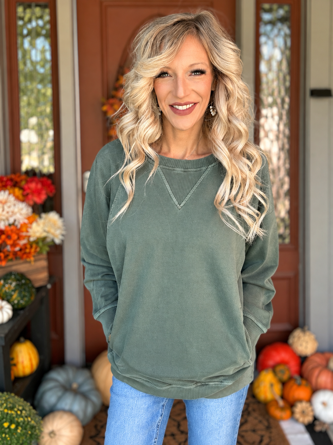 Woman wearing a sage green French terry pullover with ribbed neckline, long sleeves, side pockets, and blue jeans.
