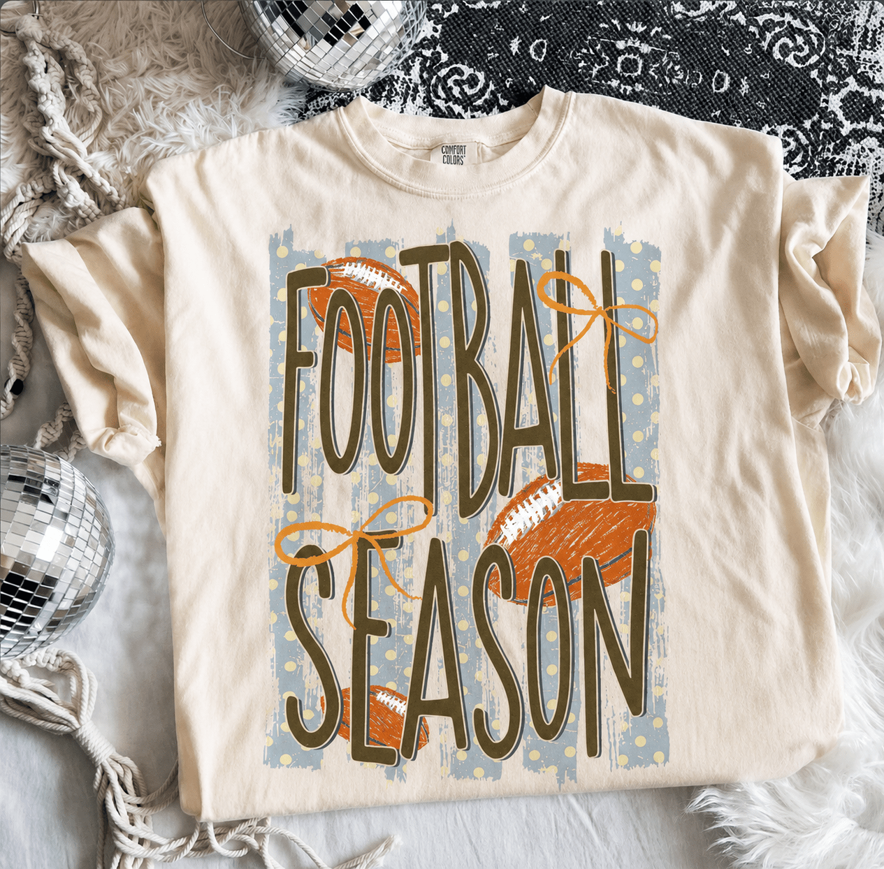 Cream Comfort Colors® Football Season graphic tee featuring vintage football artwork, distressed lettering, orange bows, and a relaxed unisex fit.