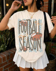 Cream Football Season graphic tee with blue stripes, brown footballs, orange bows, and playful polka dot details.