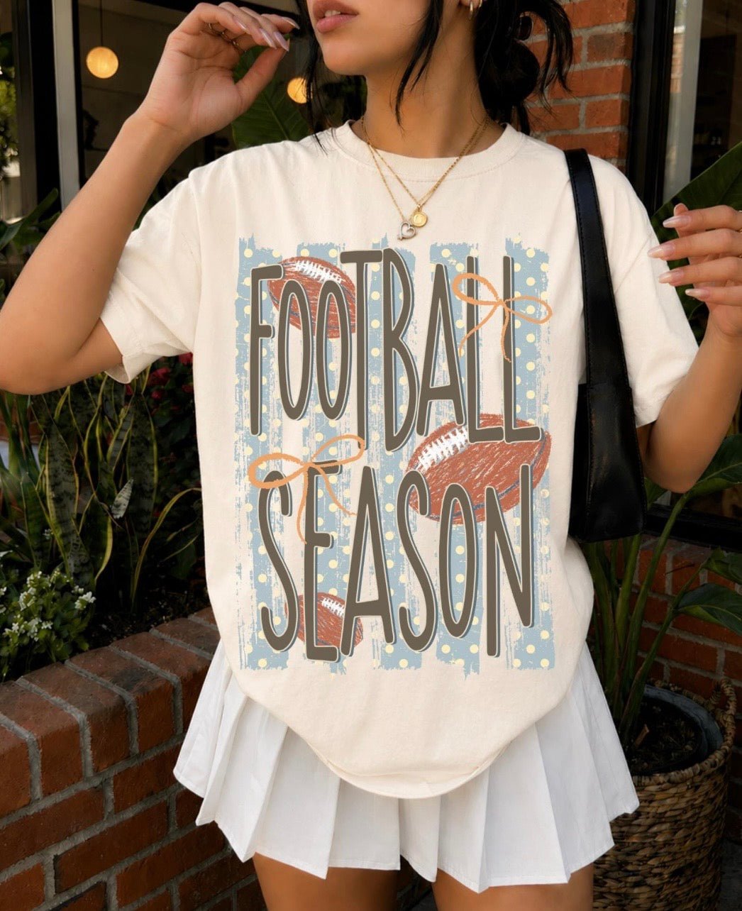 Cream Football Season graphic tee with blue stripes, brown footballs, orange bows, and playful polka dot details.