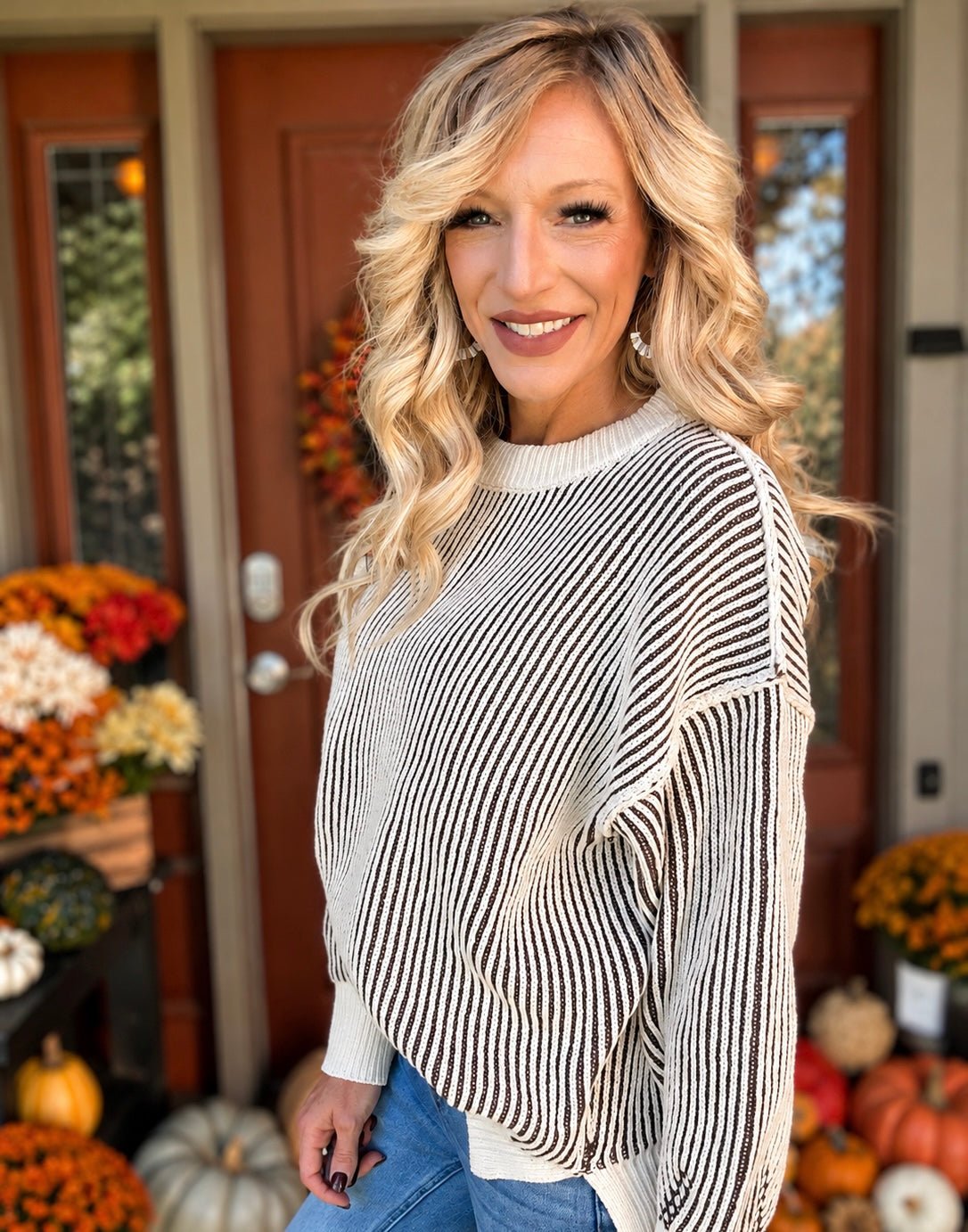 A smiling blonde woman wearing a cream and black vertically striped knit sweater with wide draped sleeves, standing in front of a decorated autumn front porch with pumpkins and fall flowers.