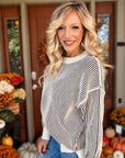 A smiling blonde woman wearing a cream and black vertically striped knit sweater with wide draped sleeves, standing in front of a decorated autumn front porch with pumpkins and fall flowers.