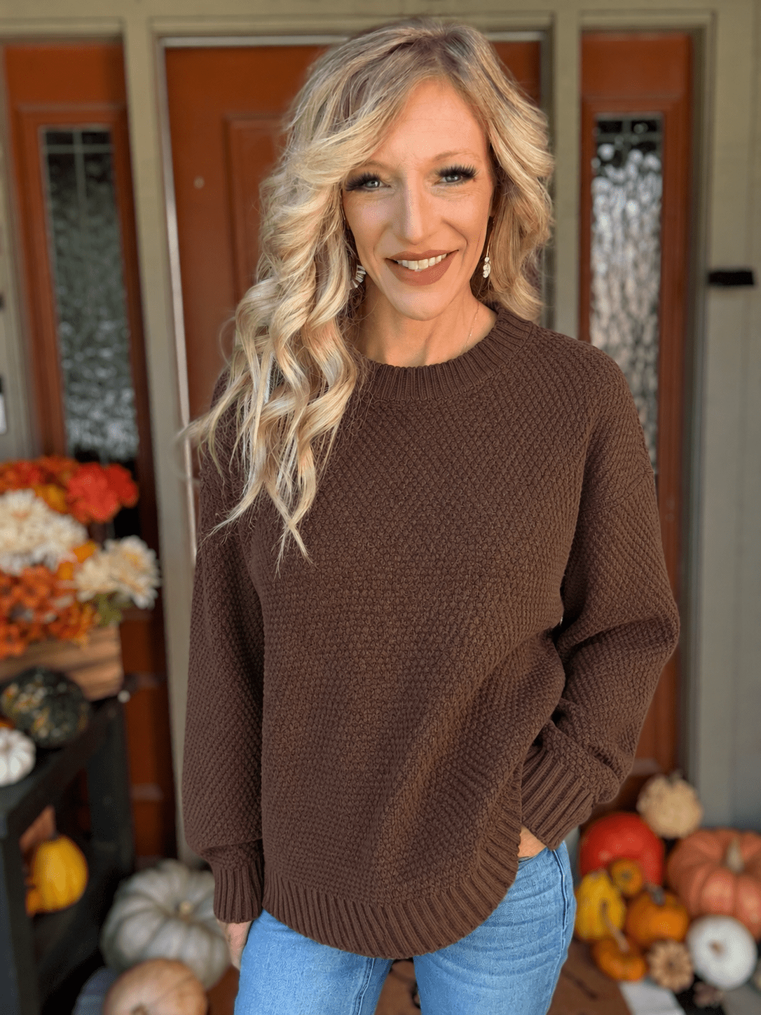 Woman wearing a chocolate brown textured knit crewneck sweater with long sleeves and ribbed cuffs, styled with blue jeans.