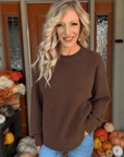 Woman wearing a chocolate brown textured knit crewneck sweater with long sleeves and ribbed cuffs, styled with blue jeans.