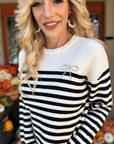 Close-up of a black and white ribbed knit sweater showing horizontal stripes, a white crew neckline, and a pearl-embellished bow on the chest.
