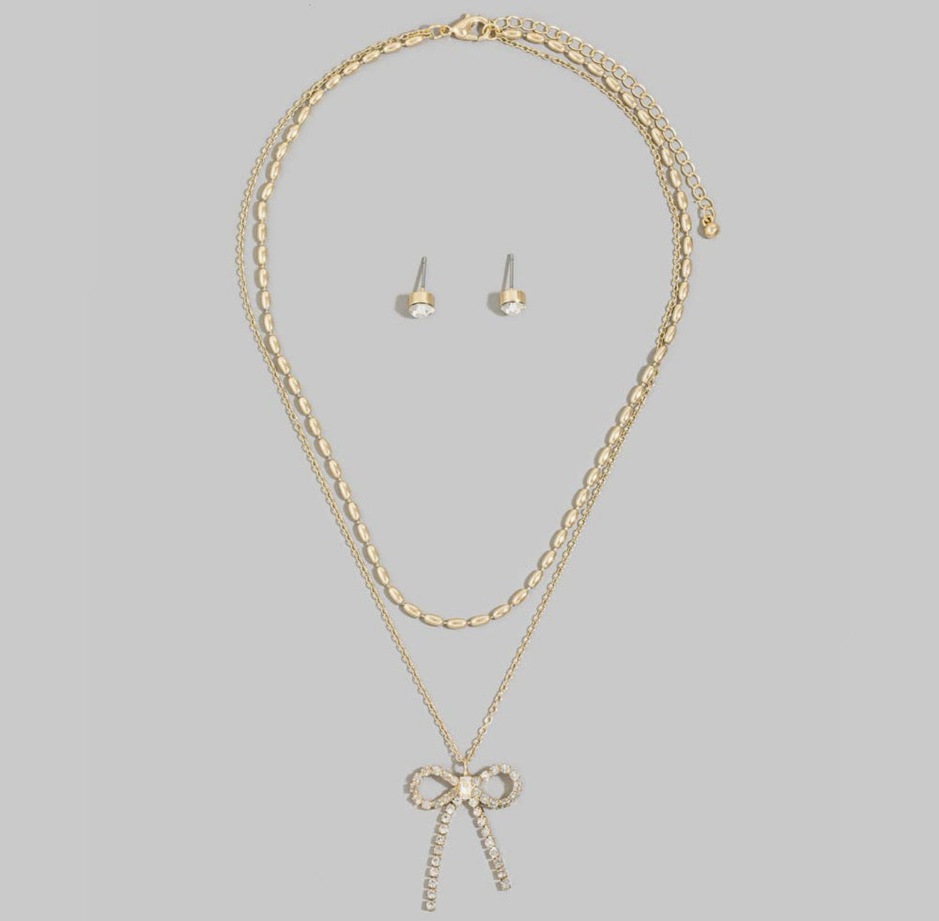 Layered gold necklace featuring an oval bead chain, crystal bow pendant, matching crystal stud earrings, and adjustable extender.