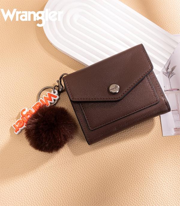 A brown textured leather Wrangler key case with a flap closure and silver snap button, shown with a dark brown pom-pom keychain and orange letter charm on a beige textured surface.