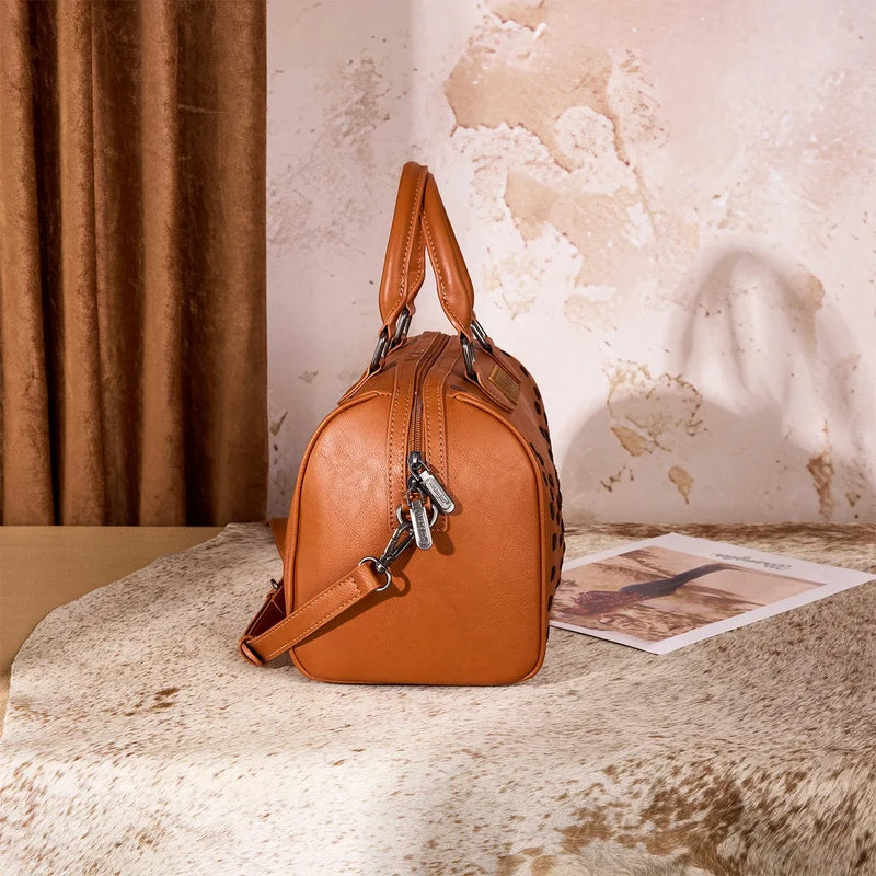A brown leather handbag with a top handle and shoulder strap, styled on a textured hide rug next to an open magazine against a rustic wall and curtain backdrop.